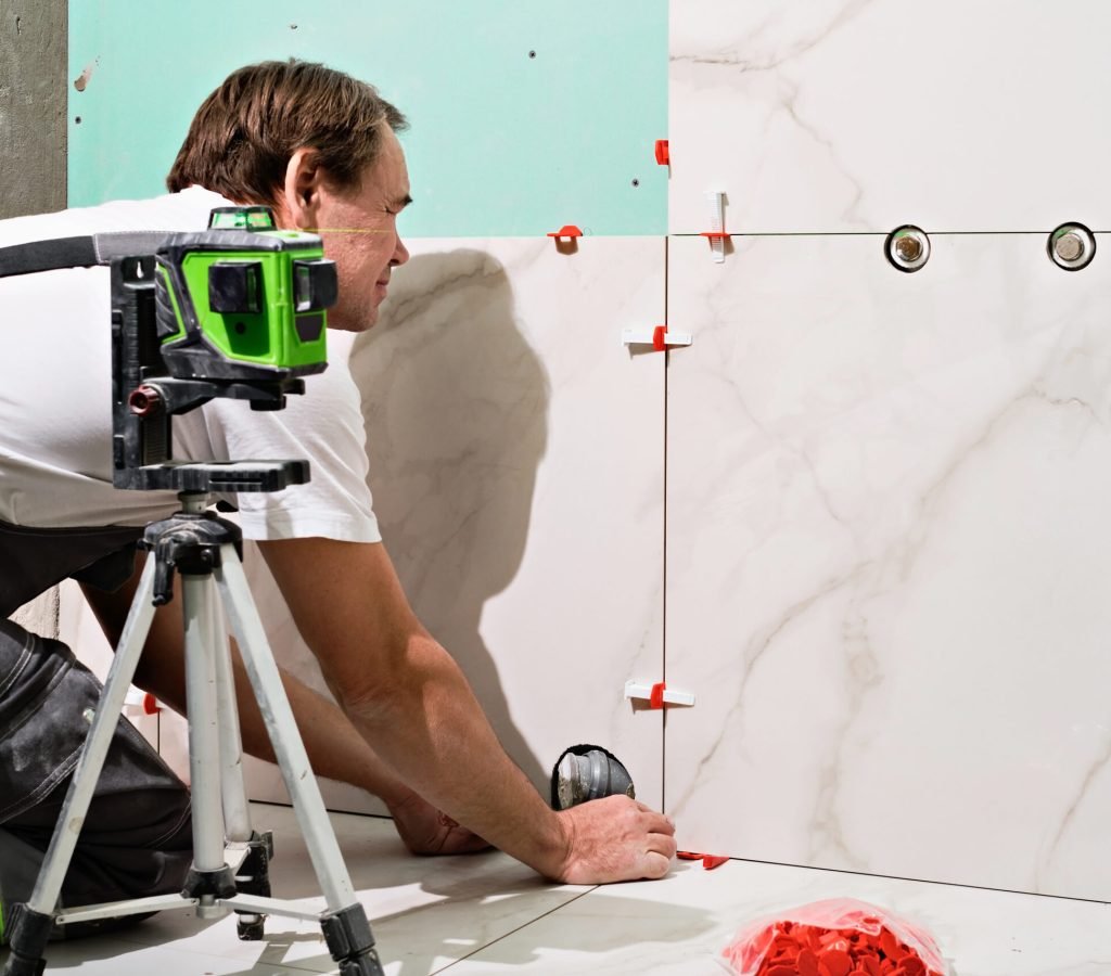 Professional master man laying ceramic tiles on wall in bathroom. Portrait of an experienced repairman laying large size porcelain tiles.Construction worker checking tile installation with laser level